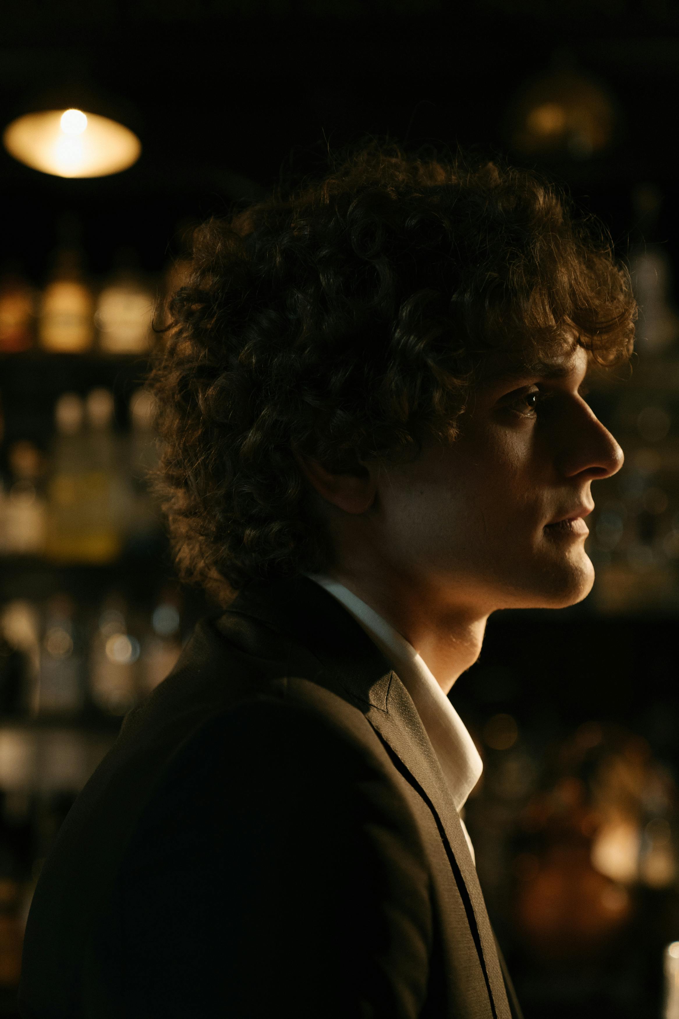 Profile of an elegant young man with curly hair sitting in a dimly lit bar.