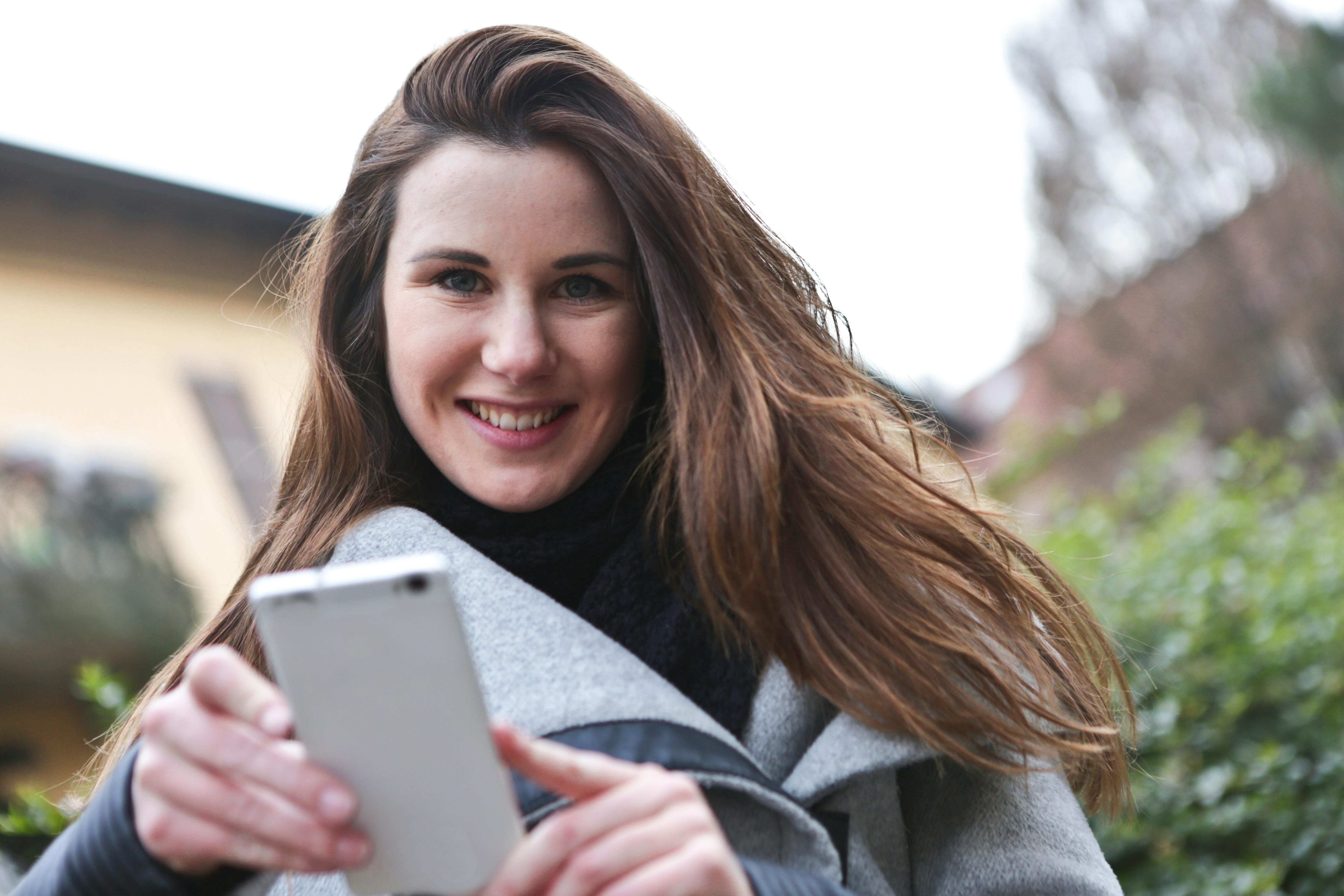 A young woman happily uses her smartphone outside, capturing a casual moment.